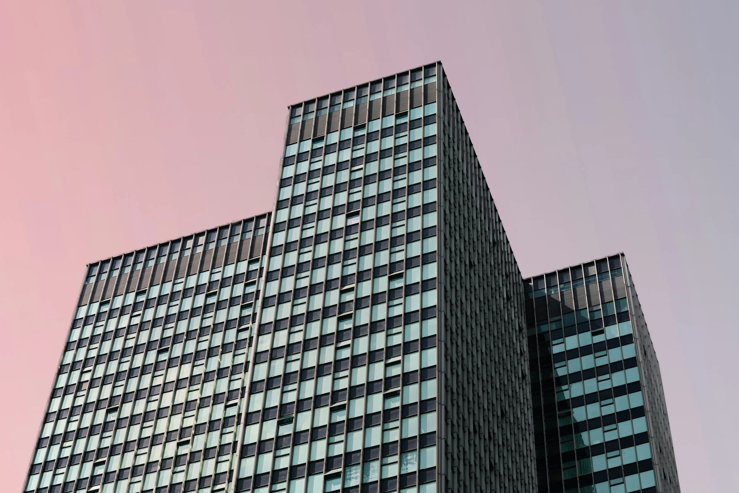 Modern high-rise office building with glass windows against a pastel pink and gray sky.