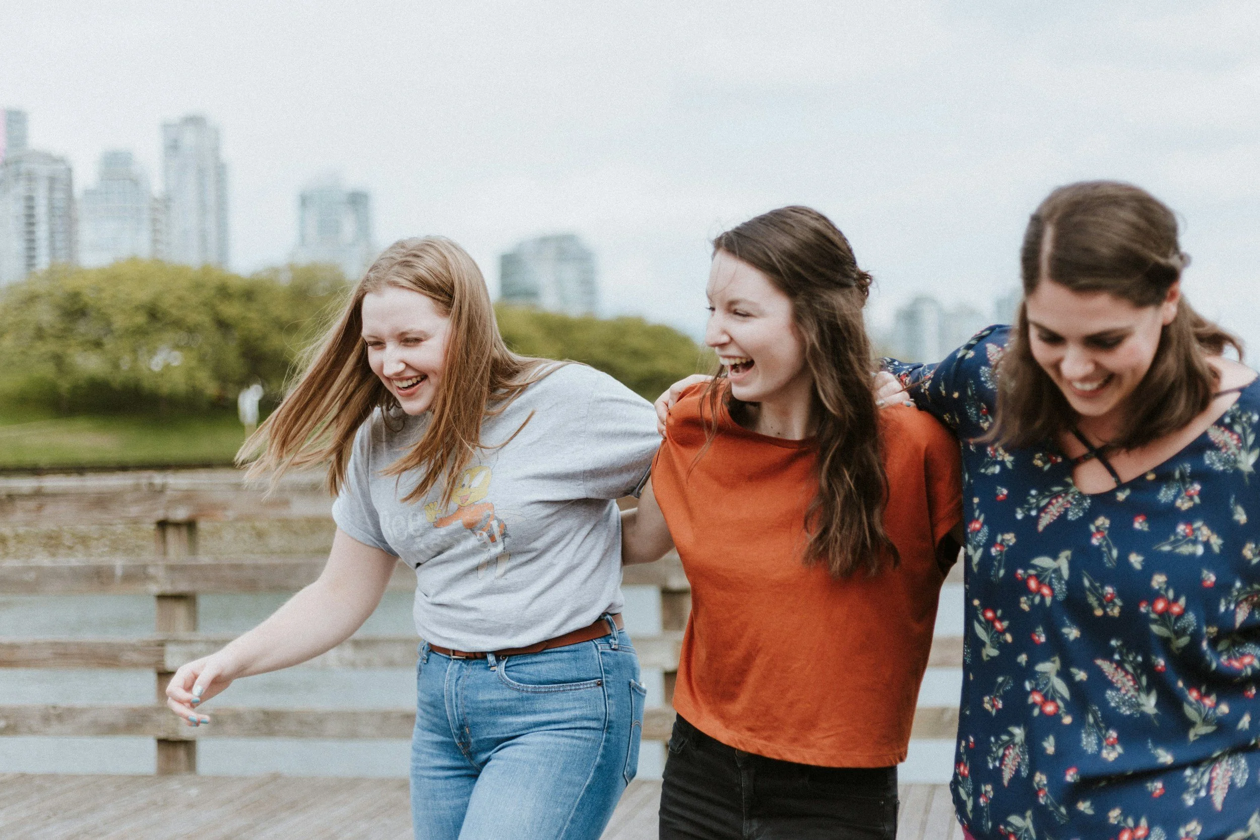 Three young women smiling and holding onto each other as they walk outdoors on a cloudy day with city buildings in the background.