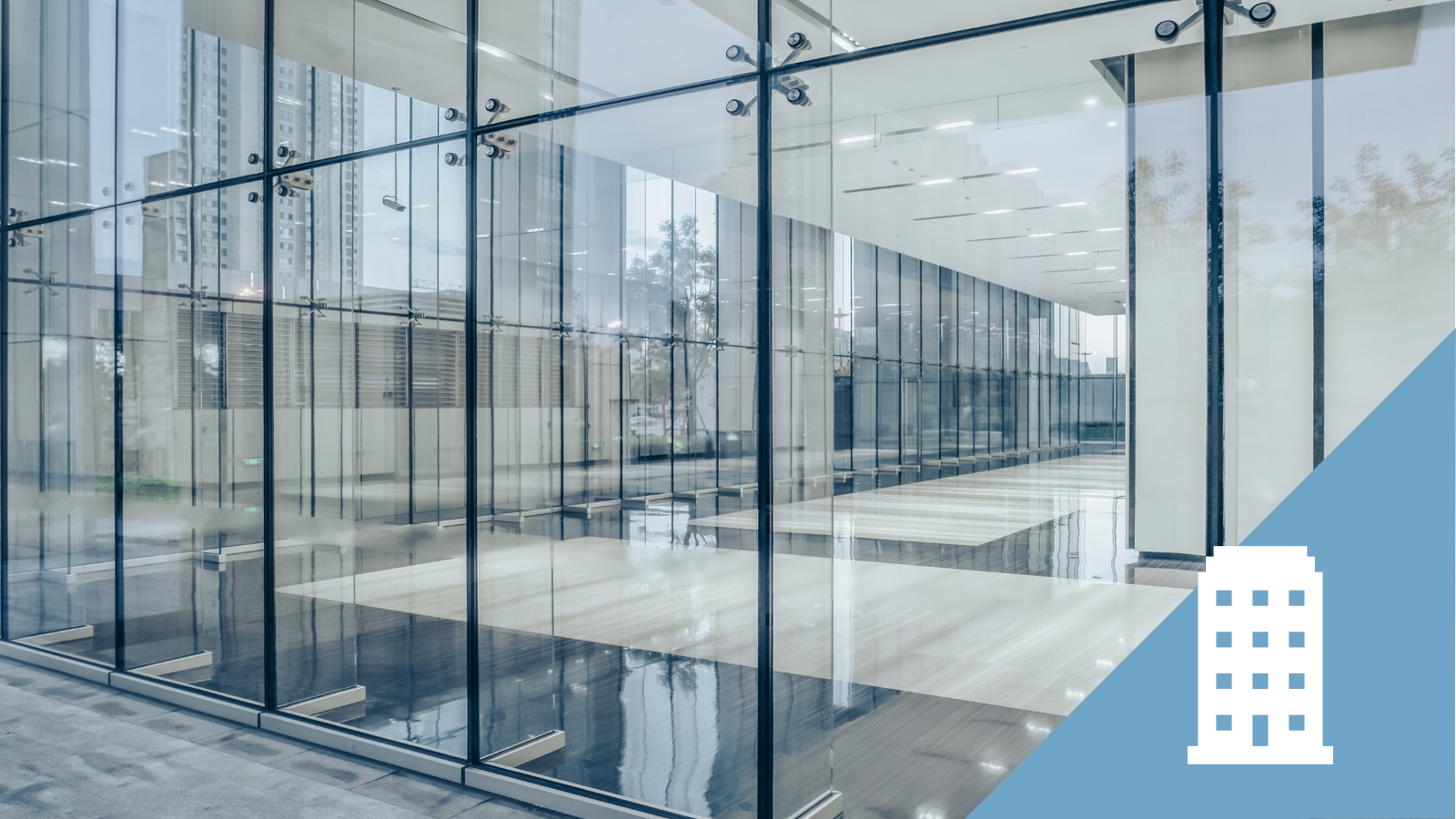 Modern building lobby with floor-to-ceiling glass walls, high ceilings, and polished floors, with reflections of trees and buildings outside.