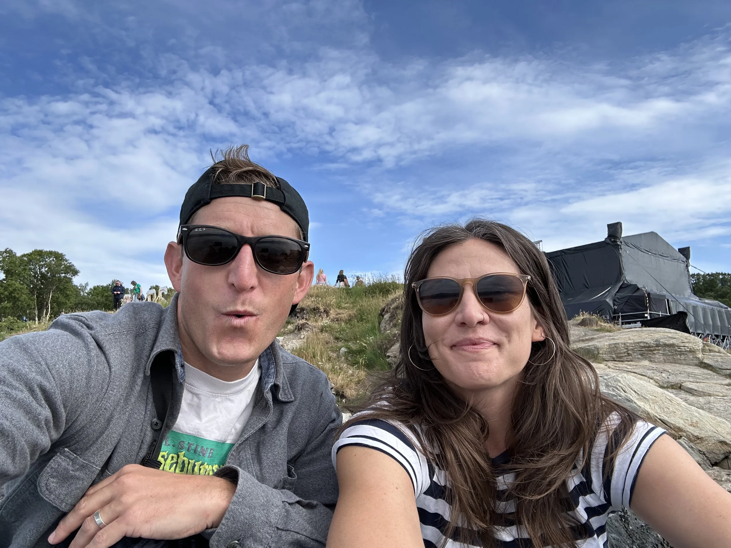 A man and woman wearing sunglasses take a selfie outdoors on a rocky hillside with a grassy field and groups of people in the background, under a partly cloudy sky.