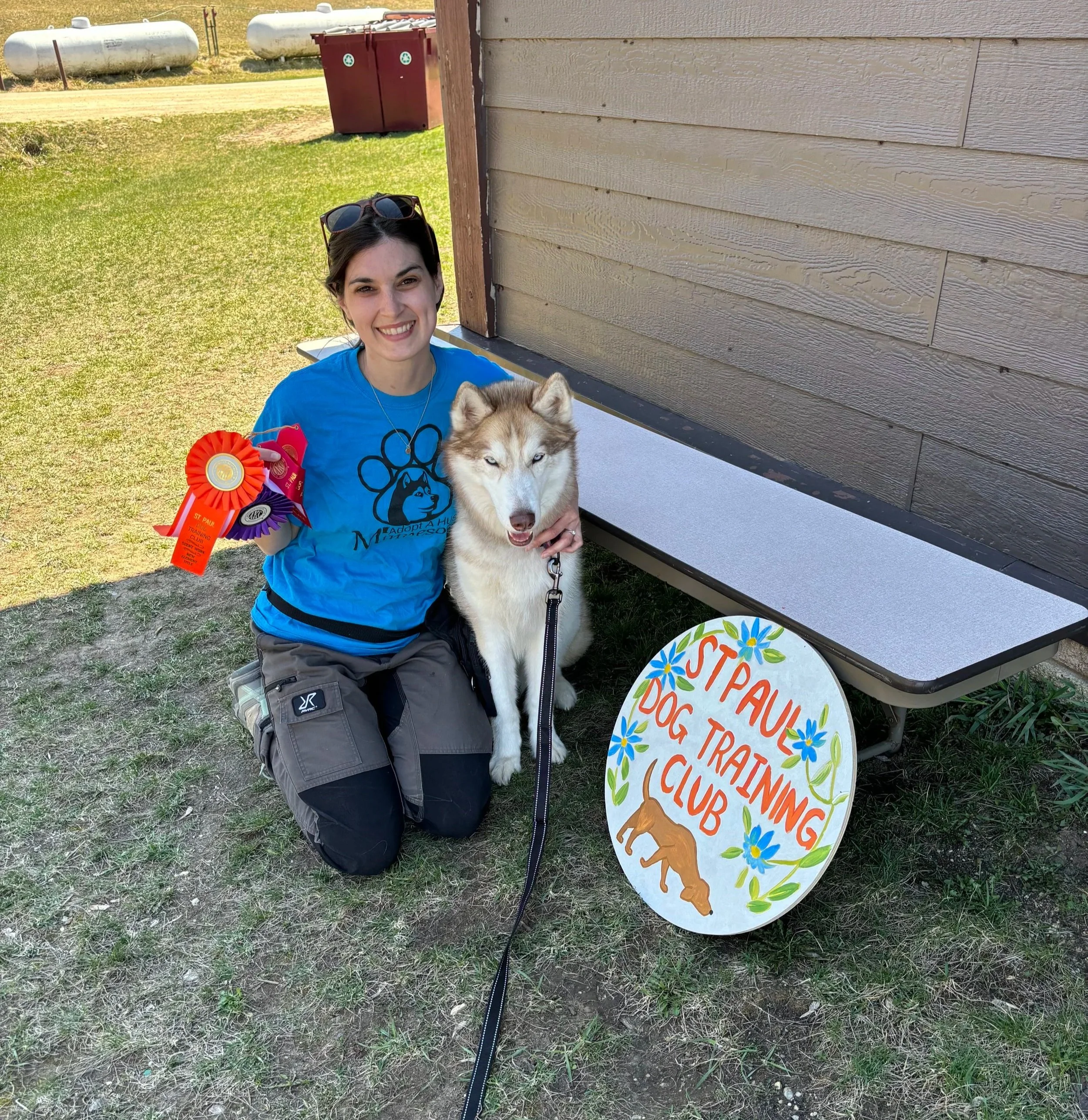 A woman kneeling next to a Siberian Husky dog holding colorful ribbons, in front of a hand-painted sign that reads "St. Paul Dog Training Club" with flowers and a dog illustration, outdoors near a building.