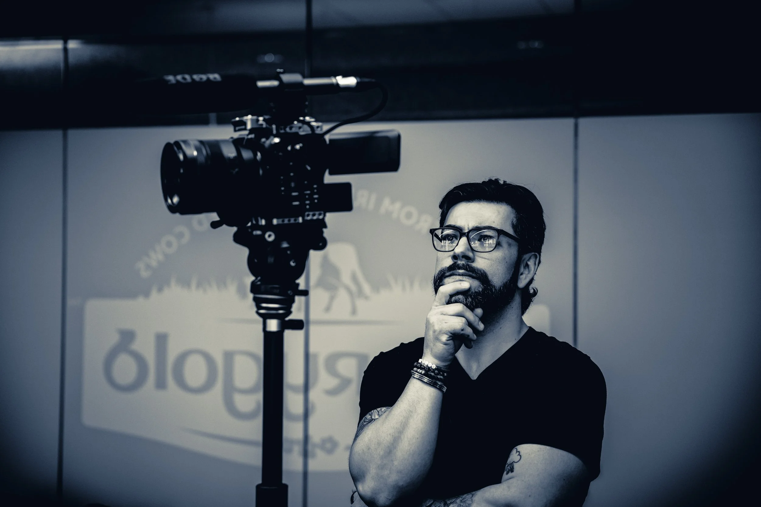 A man with glasses and a beard is standing in front of a camera on a tripod, with his hand on his chin, appearing to be thinking. The background has a logo with text and an image of a dog.