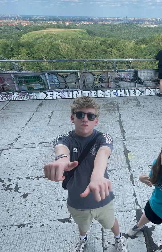 Young man with sunglasses and a black Adidas shirt pointing towards the camera on a concrete platform with graffiti and a scenic city view in the background.