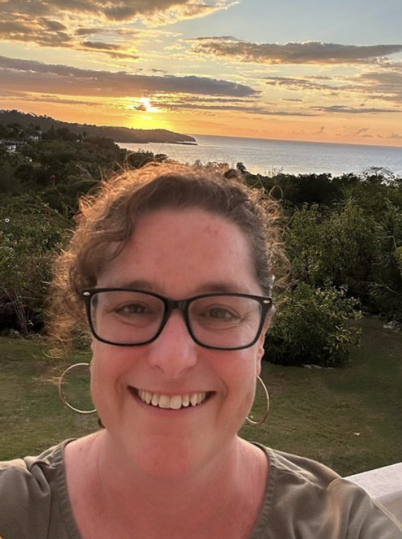 A smiling woman with glasses and hoop earrings taking a selfie at sunset, with a scenic background of greenery, water, and a colorful sky.