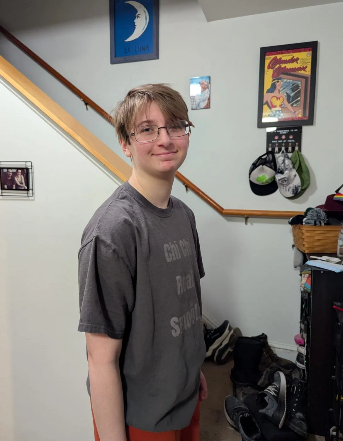 A teenage boy with glasses smiling and standing in a room with a staircase, shoes, and wall art.