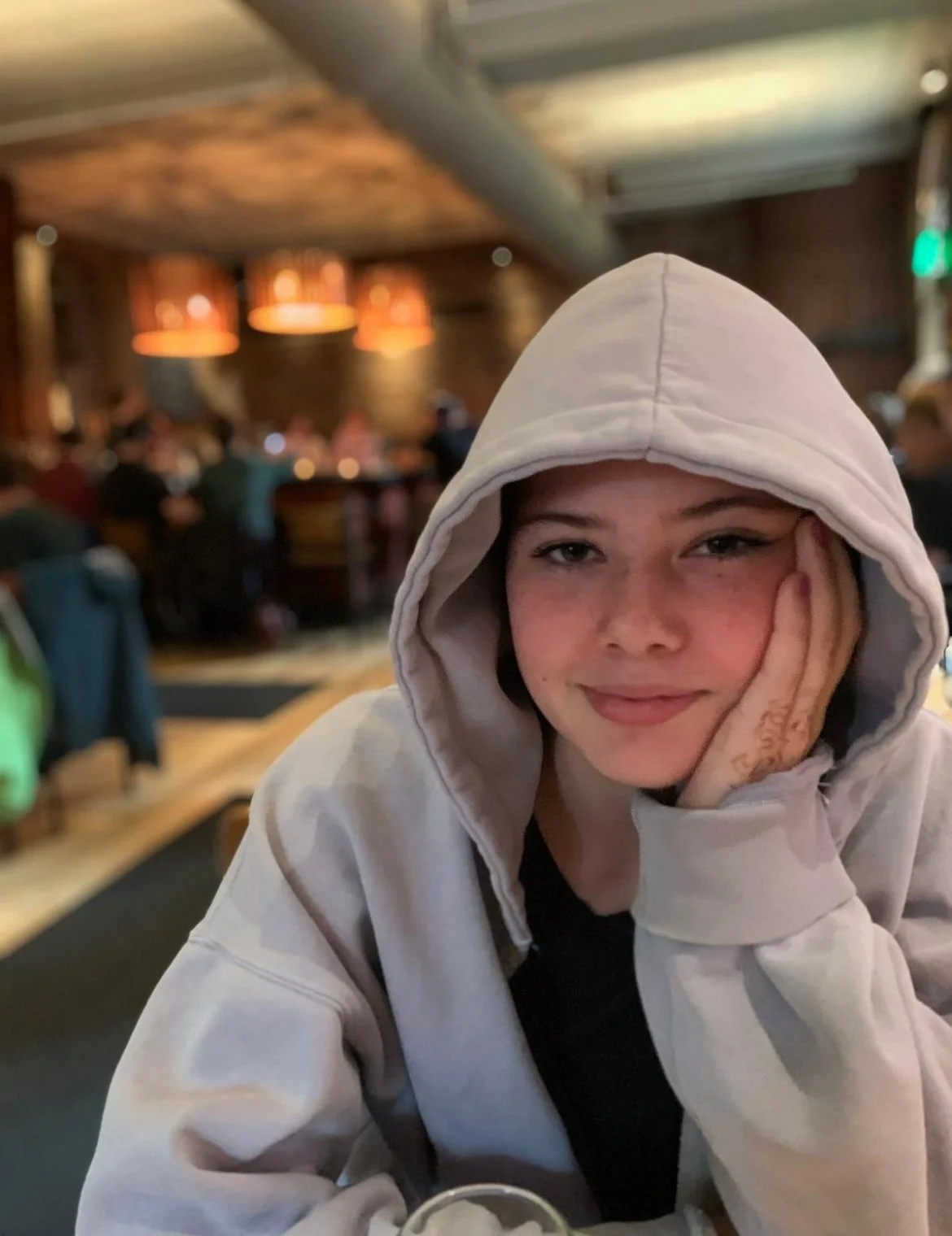 A young woman wearing a beige hoodie, smiling with her head resting on her hand, in a dimly lit indoor setting with a blurred background of other people sitting at tables.