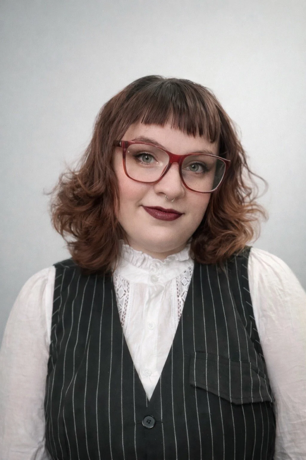 A woman with glasses and a septum piercing smiling in front of a green background, wearing a brown cardigan with yellow and white floral patterns and a silver necklace.