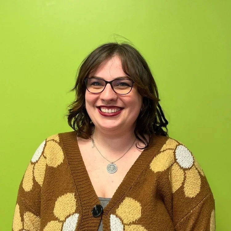 A woman with glasses and a septum piercing smiling in front of a green background, wearing a brown cardigan with yellow and white floral patterns and a silver necklace.