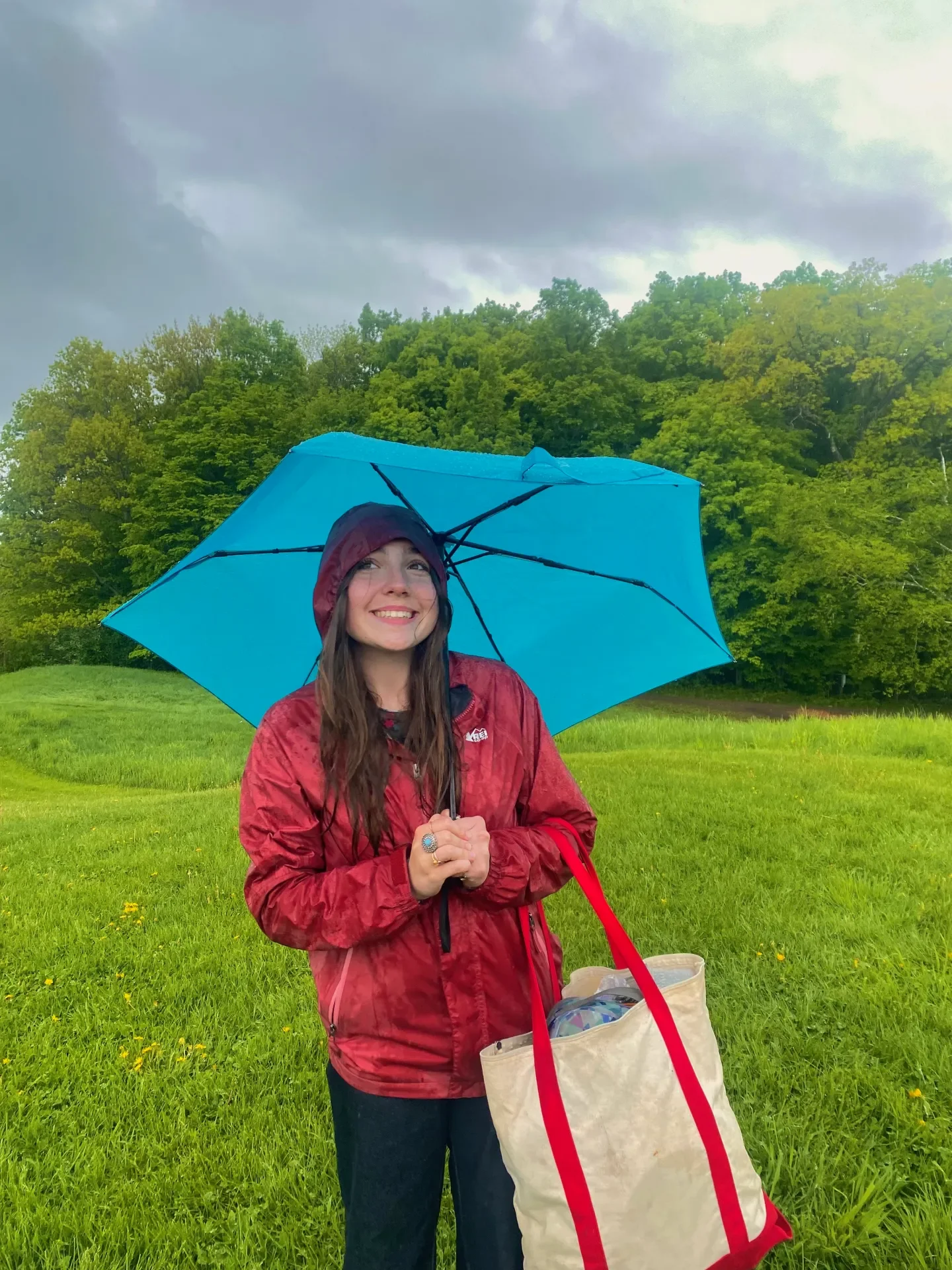 A woman standing in a grassy field holding a blue umbrella and a beige tote bag, wearing a red rain jacket and a hood, with a smiling expression, under a cloudy sky with trees in the background.