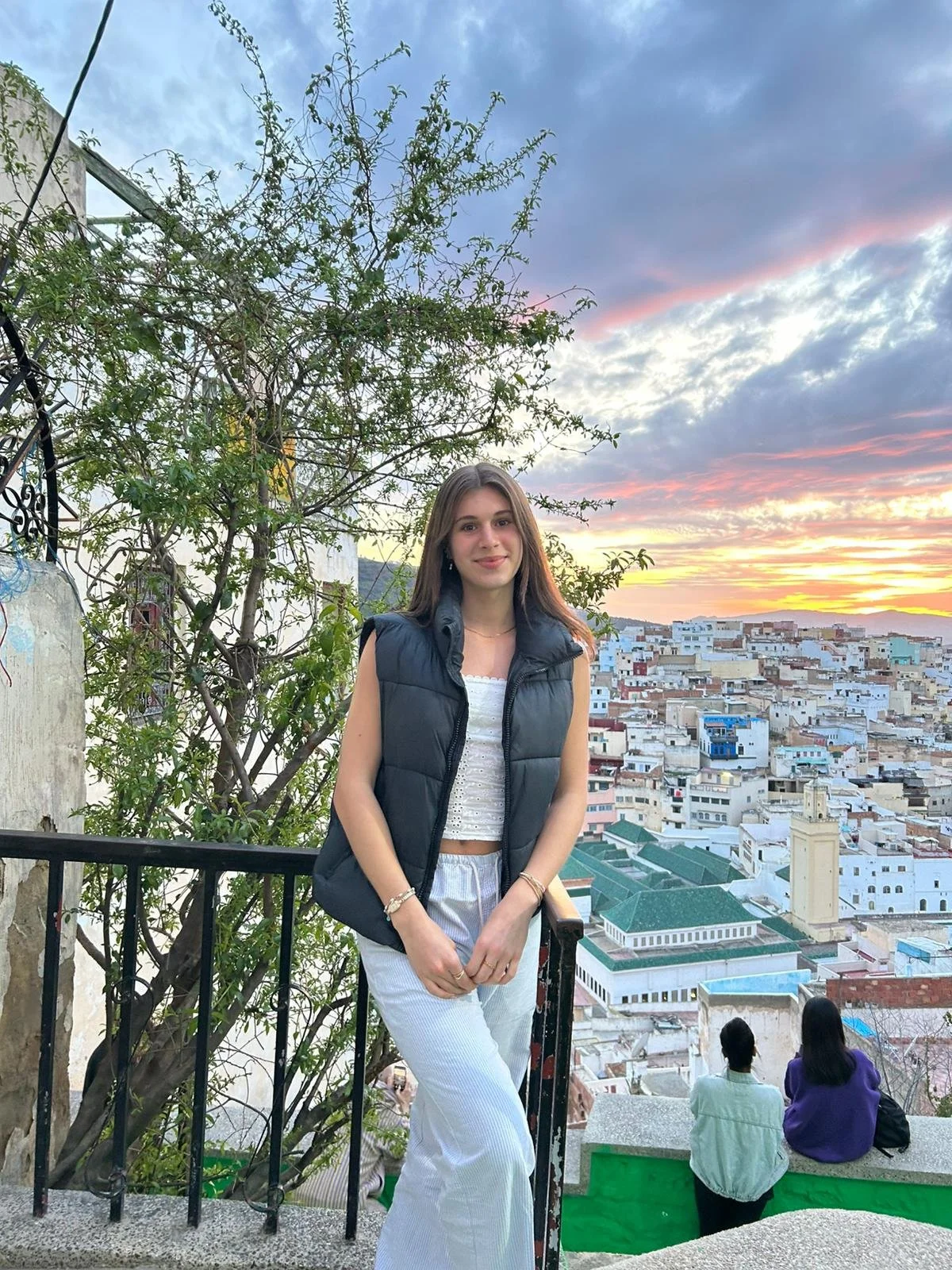 A young woman standing on a balcony overlooking a cityscape at sunset, wearing a white top, a black puffer vest, and light-colored pants, with two people sitting on a ledge in the background.