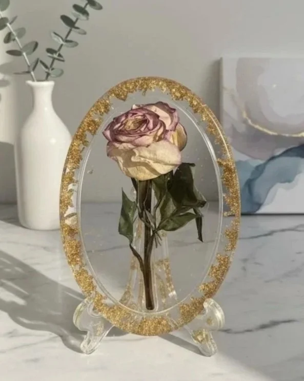 Une rose séchée dans un cadre en verre avec des paillettes dorées, posé sur un support en verre, avec une vase blanche et une plante en arrière-plan sur une surface en marbre.