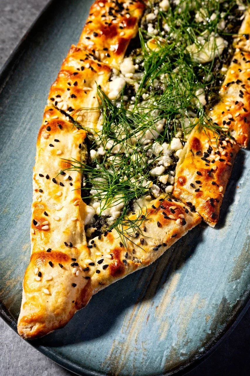 A slice of rectangular flatbread topped with cheese, sprinkled with black and white sesame seeds, garnished with fresh dill herbs, served on a dark rectangular plate.