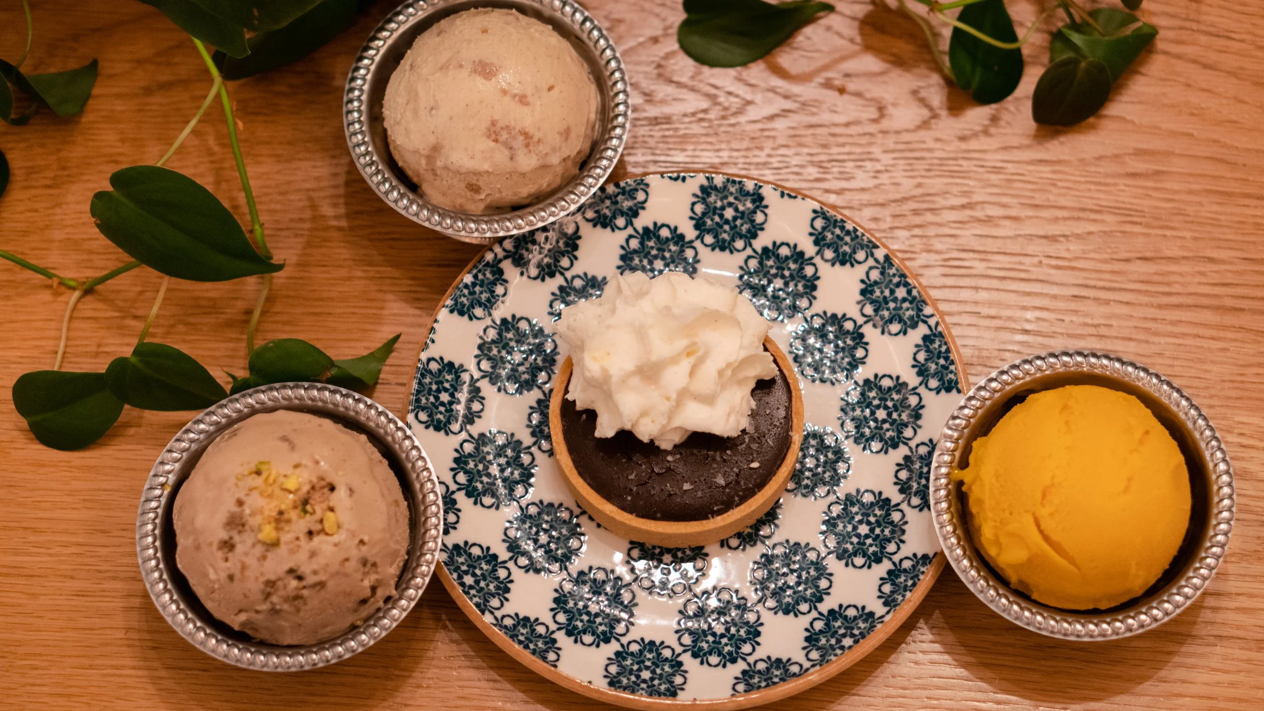 Three small metal cups with ice cream, one on a floral patterned plate with a chocolate tart topped with whipped cream, garnish of pistachio bits, and a scoop of mango sorbet on a wooden table decorated with green leaves.