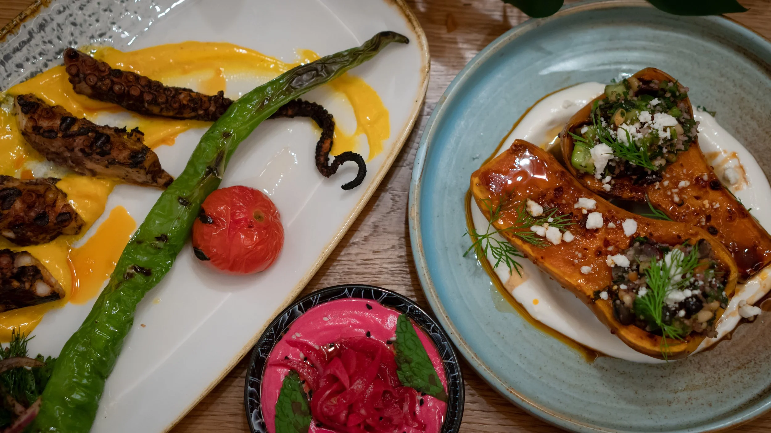 A plate with grilled octopus tentacles, a roasted cherry tomato, and a green chili pepper. Next to it, a blue bowl with stuffed poblano peppers topped with cheese and herbs. A small black dish with pickled red vegetables and mint leaves.