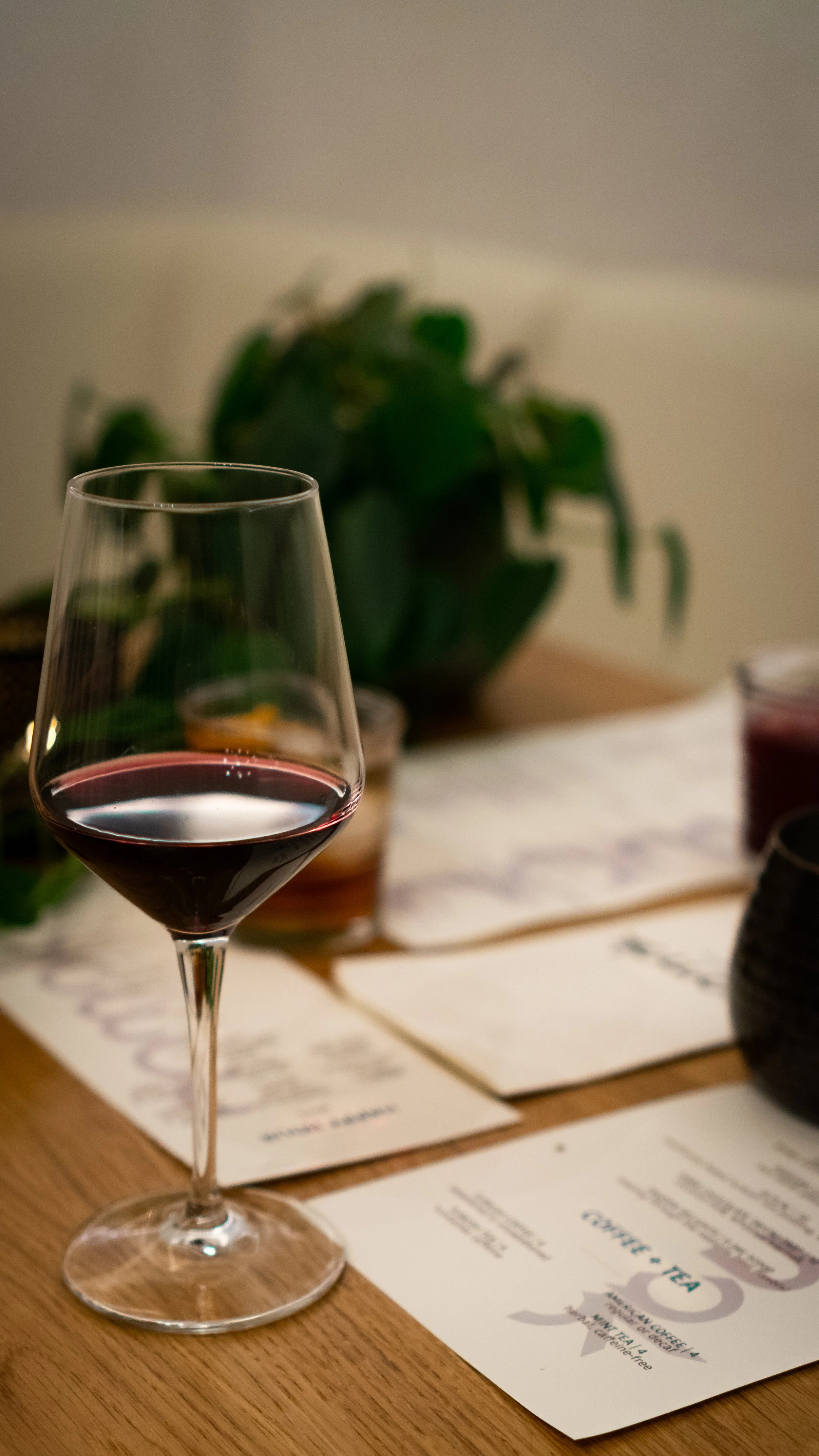 A glass of red wine on a wooden table with menus and a potted plant in the background.