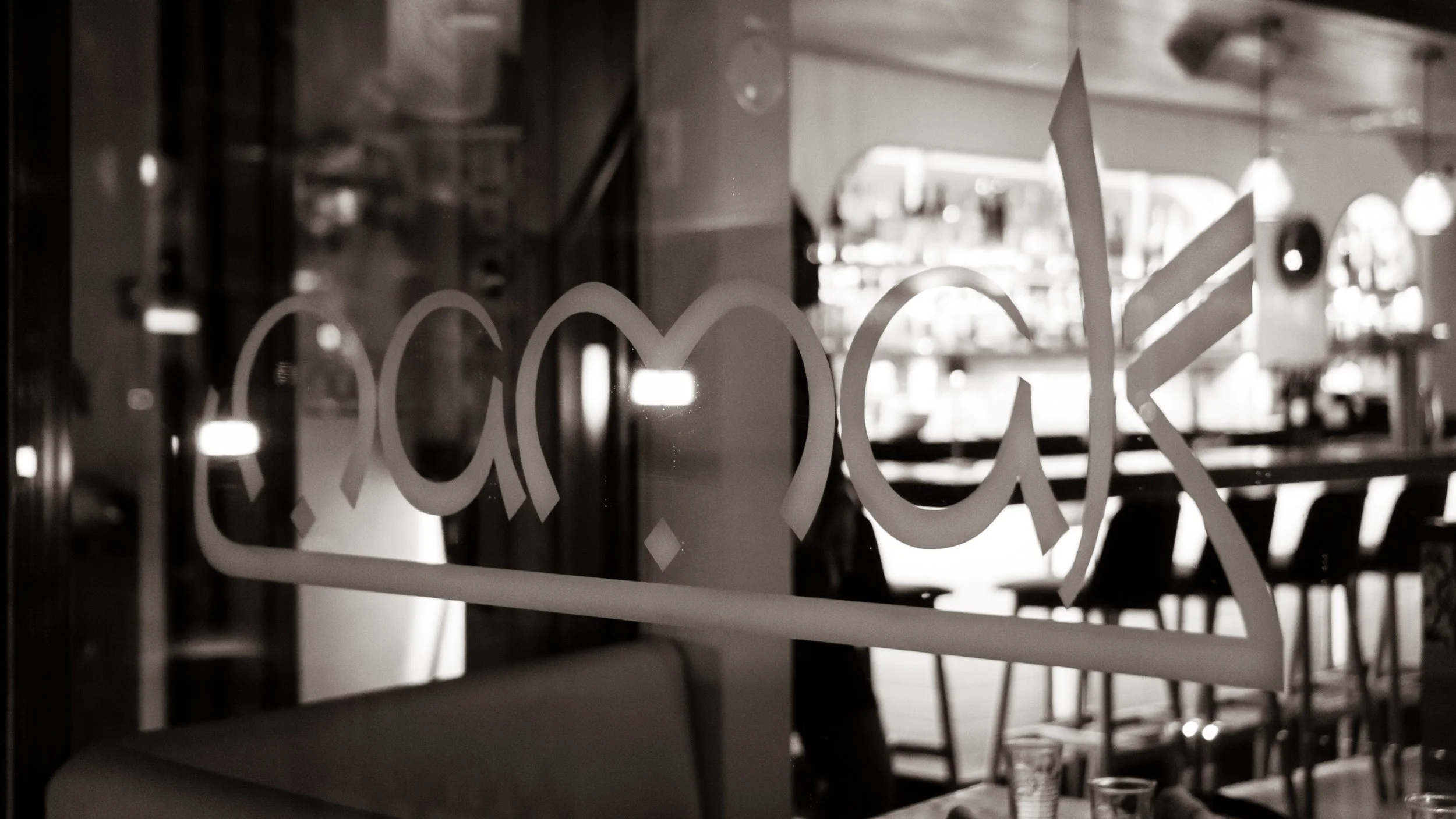 Black and white photo of a restaurant or cafe interior seen through a glass door or window with the word 'Cook' written on it.