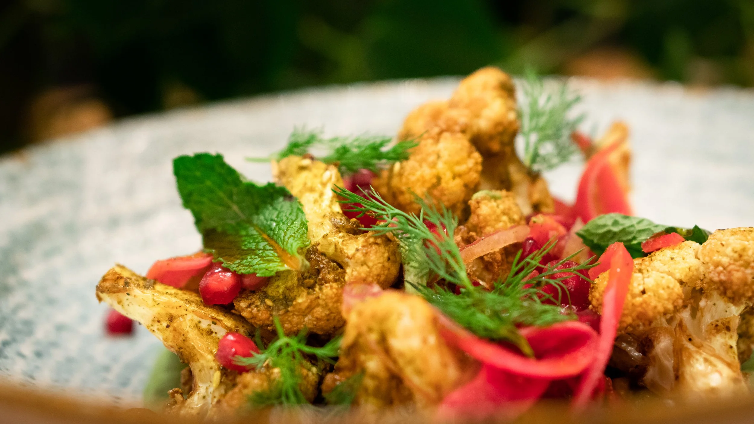 Close-up of a plated dish featuring roasted cauliflower, garnished with fresh herbs, pomegranate seeds, and sliced red onions.