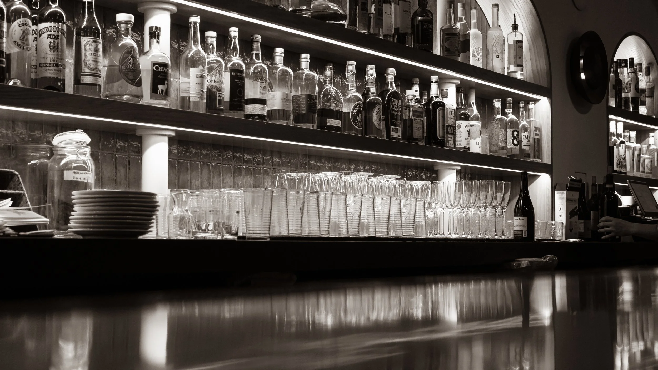 Bar with shelves of liquor bottles and glassware, including glasses and bowls, in a dimly lit setting with a reflective surface in the foreground.