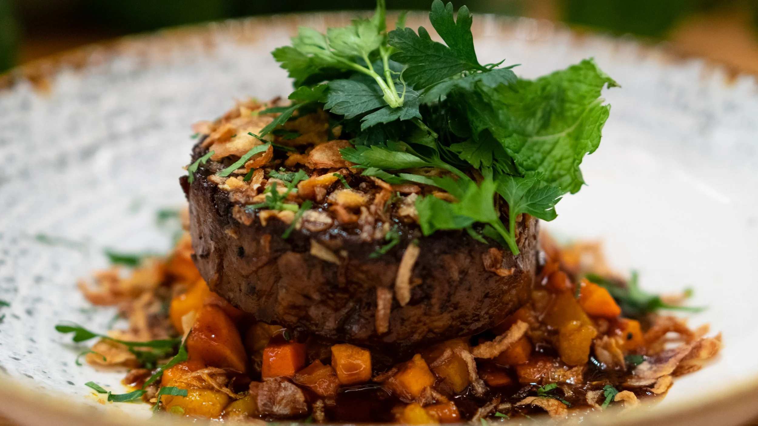 A plated gourmet beef tenderloin steak topped with fresh herbs, crispy onions, and served with sautéed vegetables and a dark sauce.