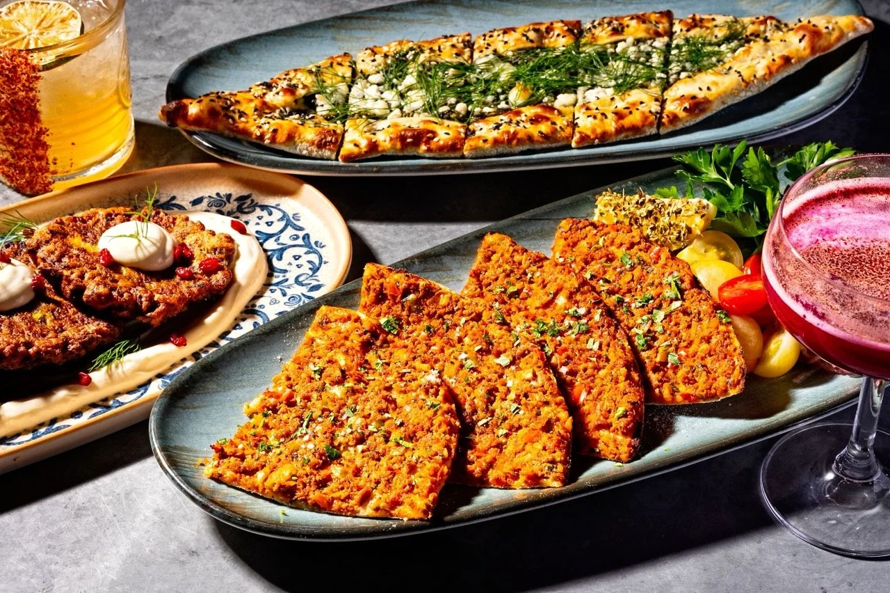Assorted dishes including slices of spicy lamb lahmacun, a flatbread garnished with herbs, zucchini fritters with sauce, and two drinks, one appearing to be an orange beverage and the other a pink cocktail.