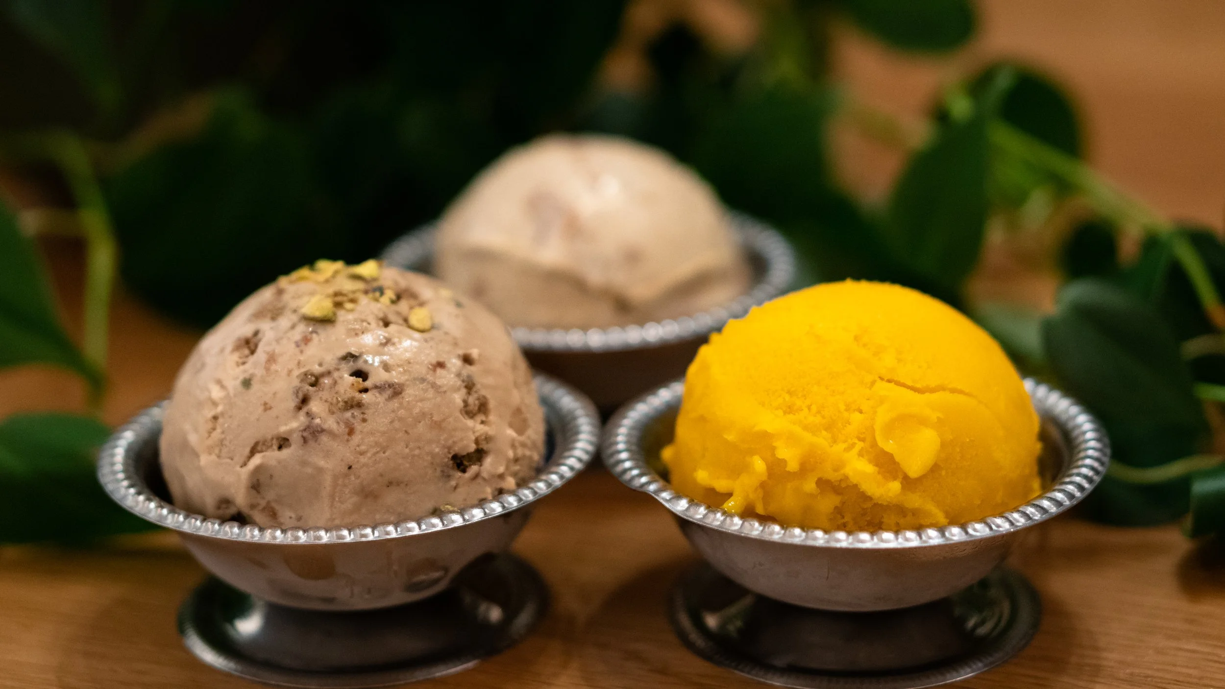 Three scoops of ice cream in small metal bowls, with green leaves around them on a wooden surface.