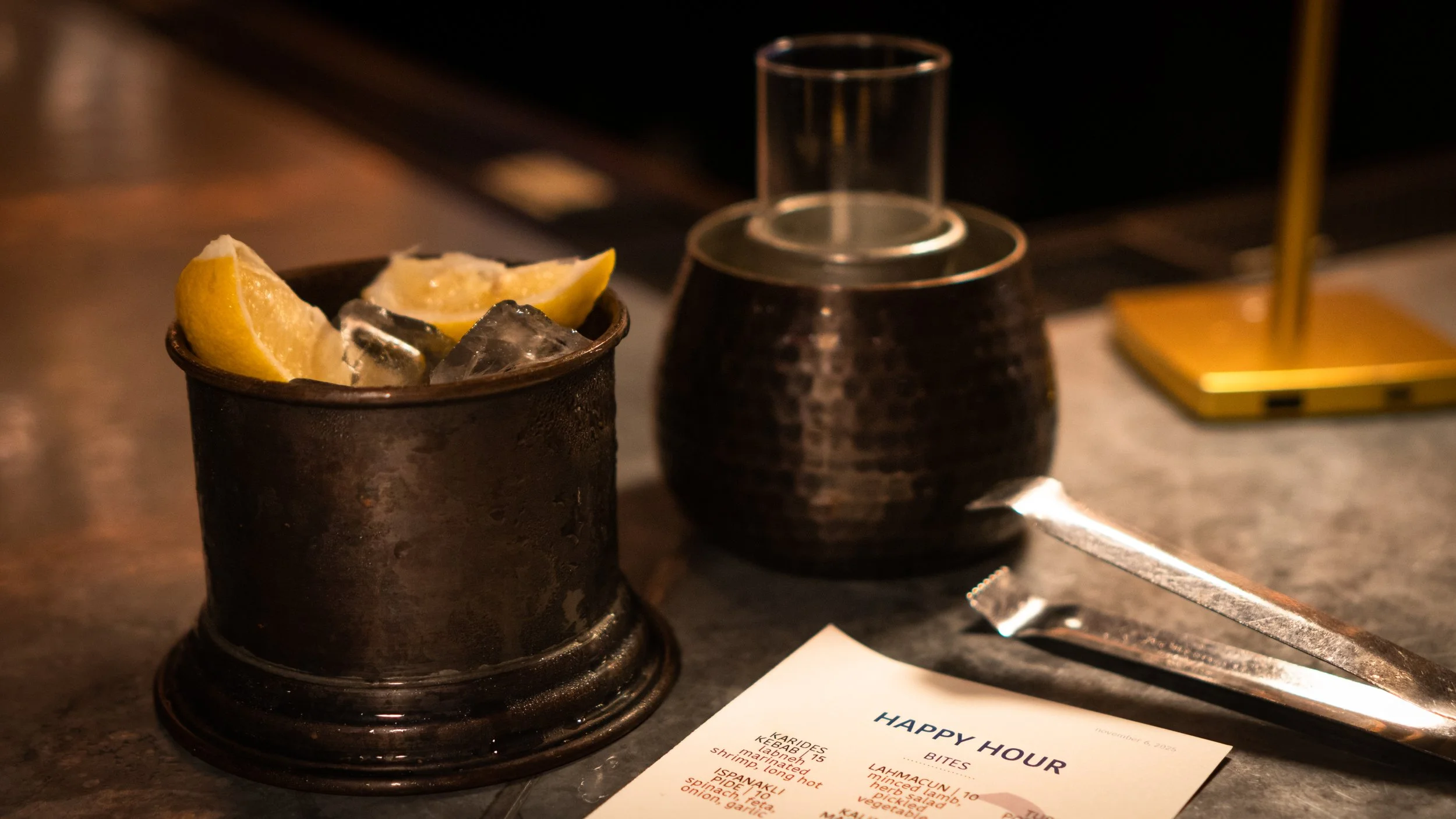 A metal container with lemon wedges and ice cubes, a dark textured pottery container with a glass on top, a pair of metal tongs, and a menu titled "Happy Hour" on a bar counter.
