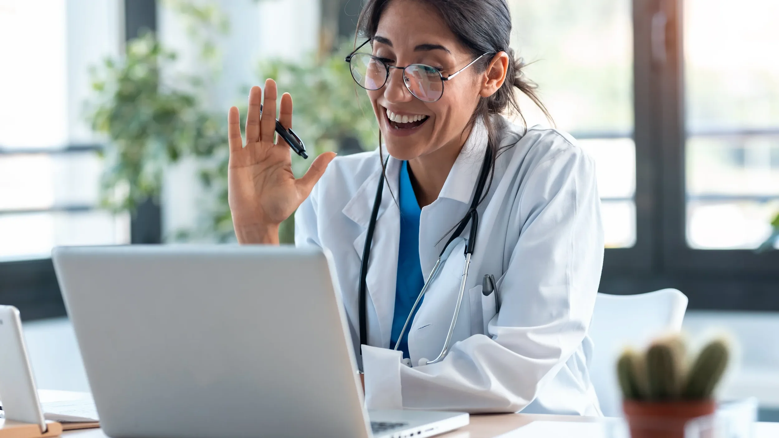 A smiling female doctor with glasses and a stethoscope waving at her laptop during a virtual patient visit.