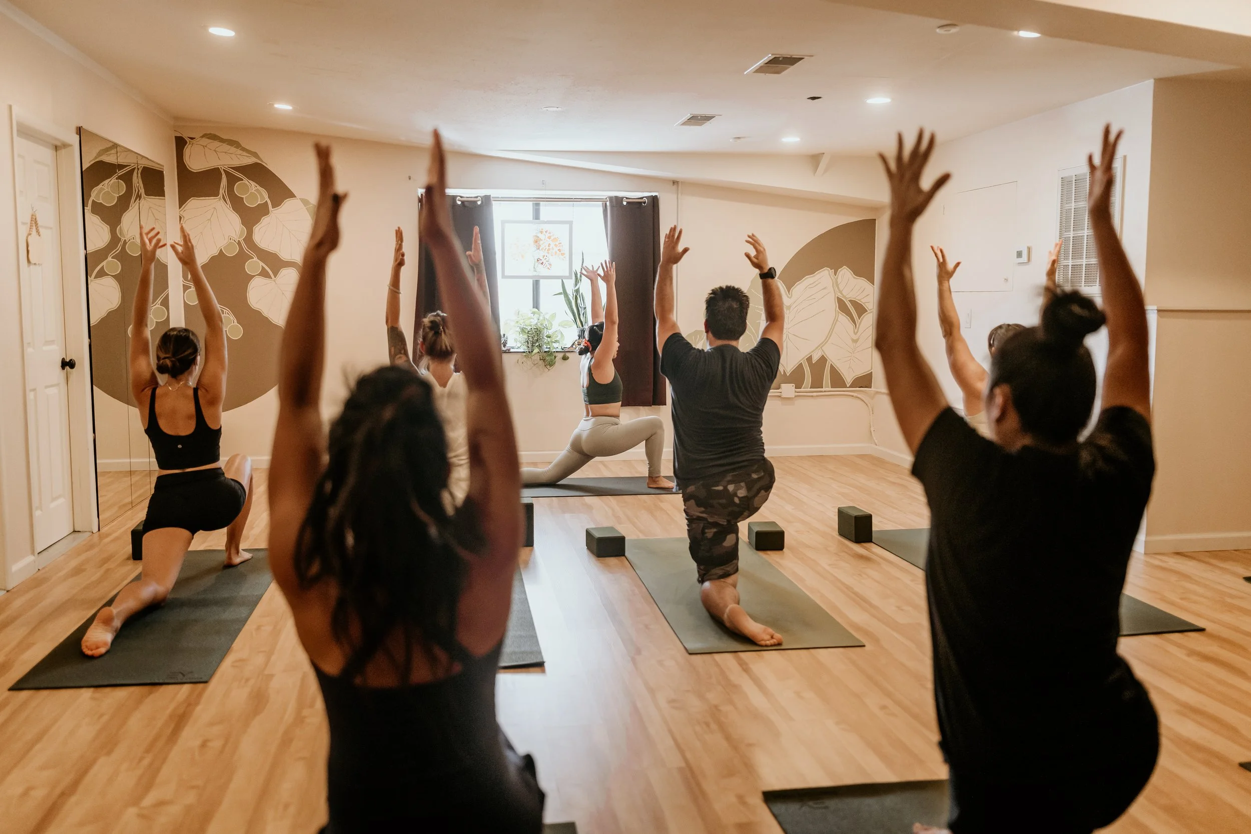 A group of people participating in a yoga class, performing a lunge pose with arms raised, in a well-lit studio with wooden floors and wall art.