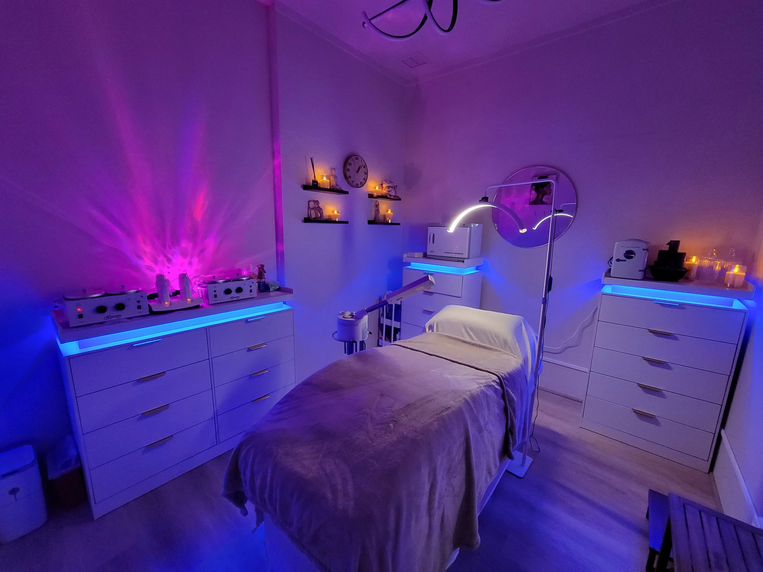 A spa or massage room with a treatment bed in the center, surrounded by white furniture with blue LED lighting, candles, and relaxing decor, creating a calming atmosphere.