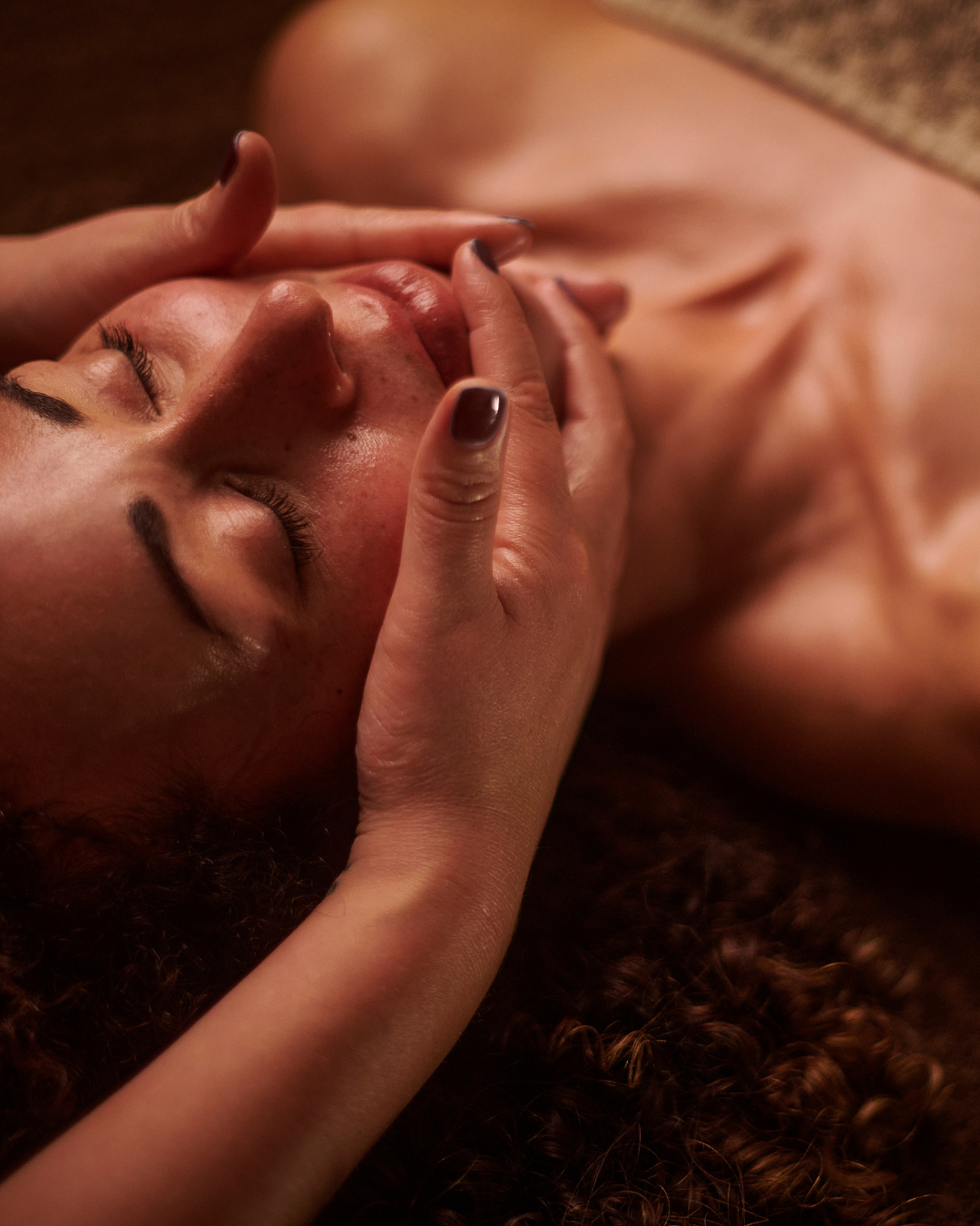 A woman with curly hair receiving a facial massage, lying down with eyes closed.