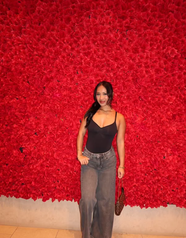 A woman standing in front of a large red textured floral wall, wearing a black sleeveless top, gray high-waisted pants, and holding a brown purse.