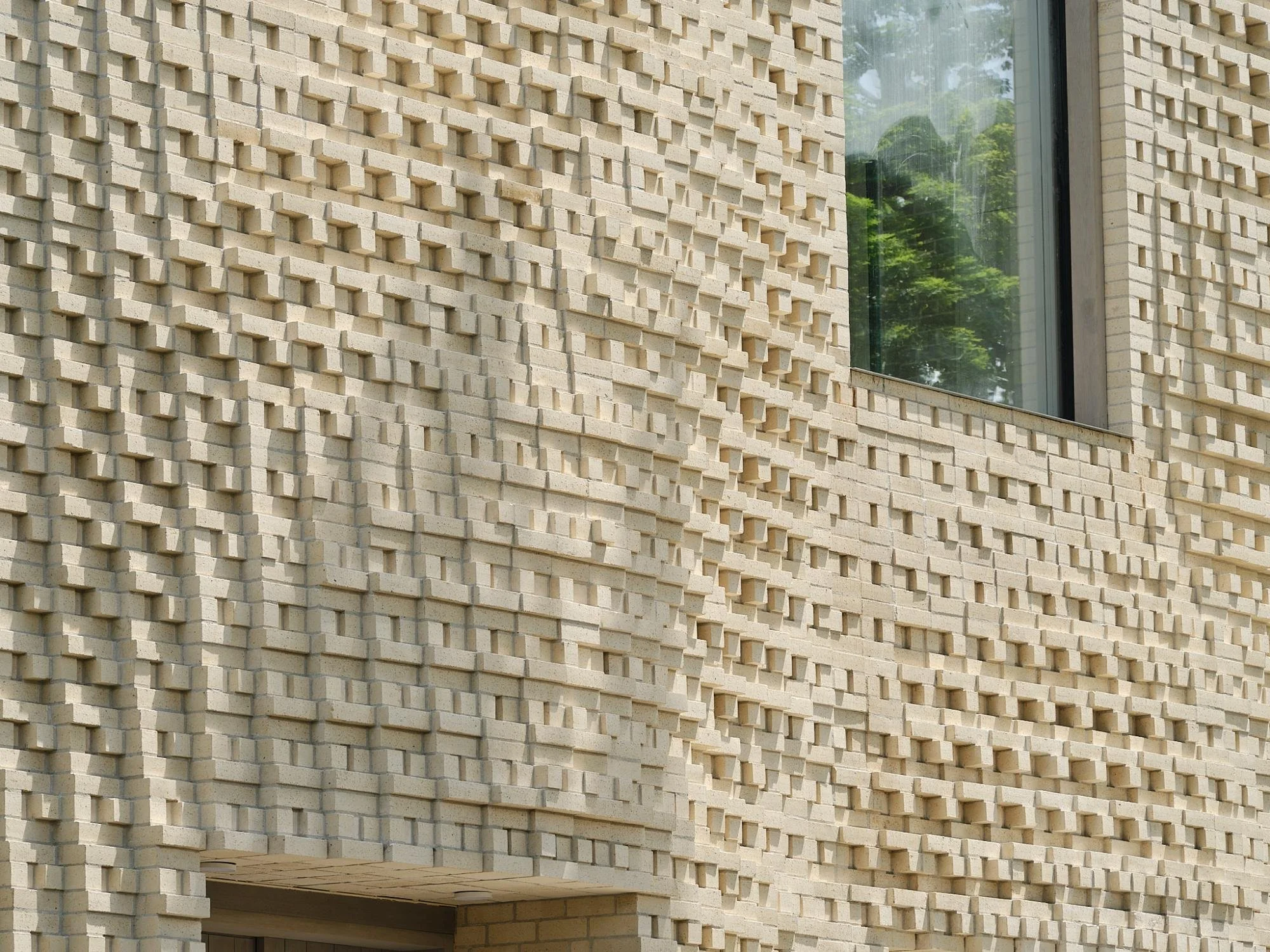 Close-up of a modern building wall made of beige bricks with a textured pattern, and a large window reflecting greenery.
