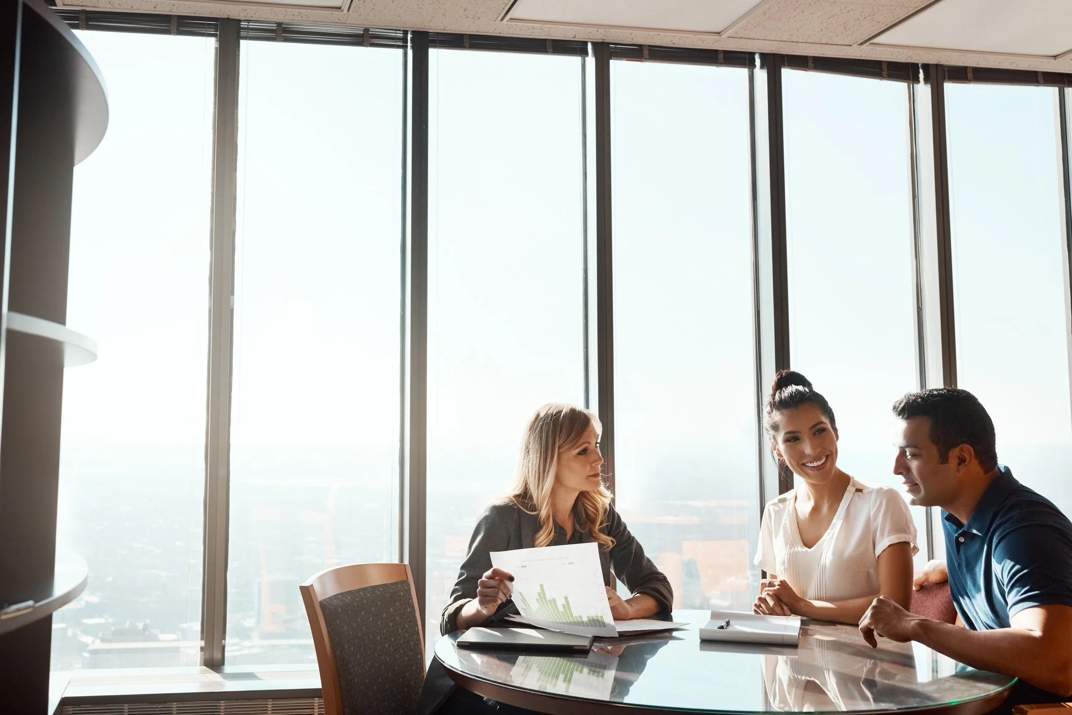 Three business professionals having a discussion at a round table in a modern office with large windows showing a cityscape in the background.