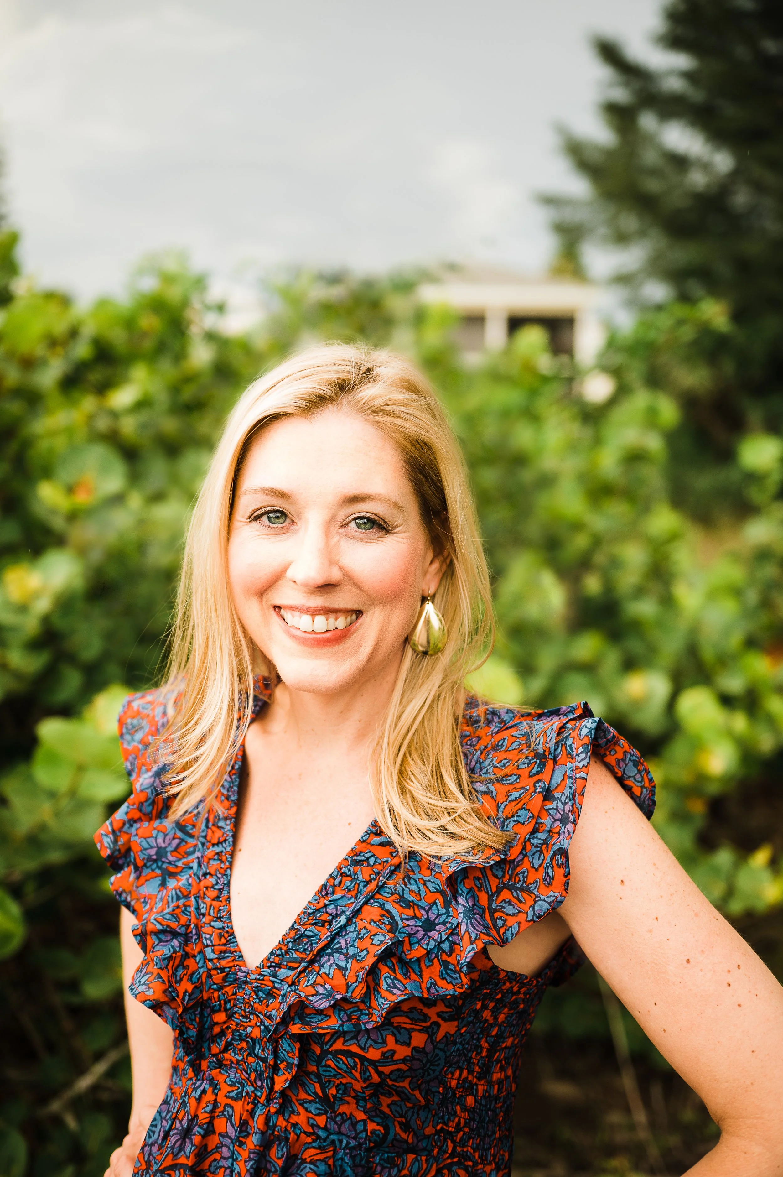 A smiling woman with blonde hair, wearing a colorful patterned dress with ruffle sleeves, standing outdoors among green foliage.