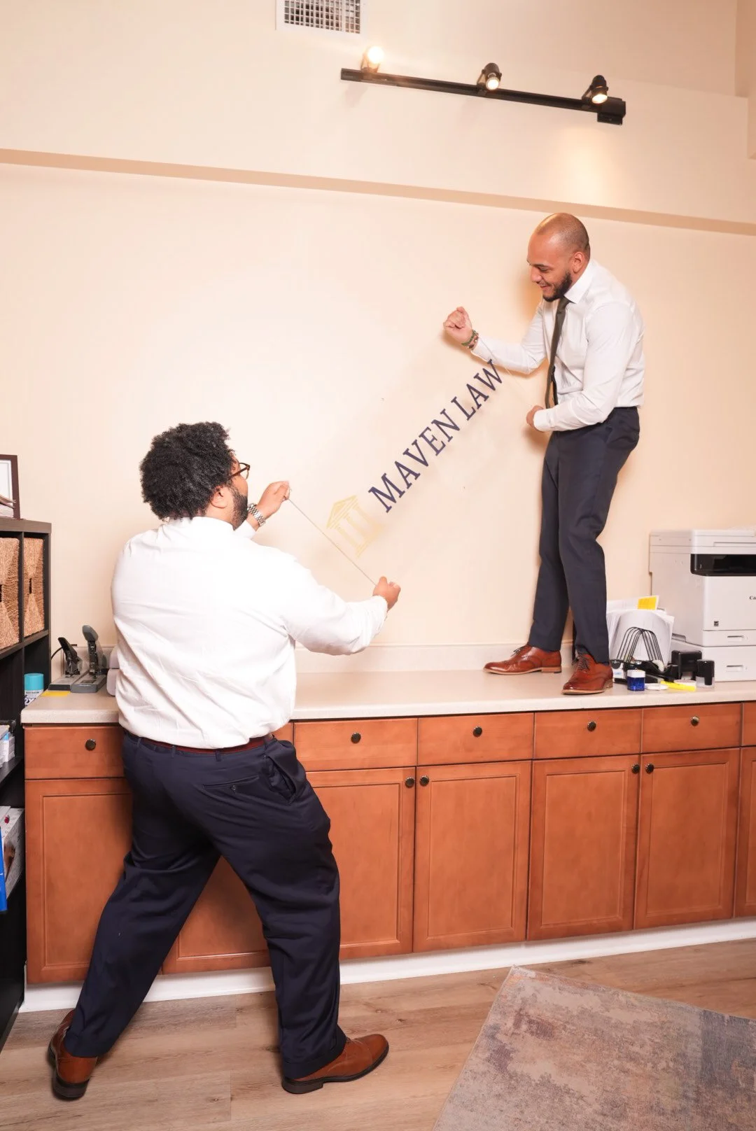 Two men in business attire playfully pulling on a rope attached to a wall sign that reads 'Maven Law.' One man is standing on a counter while the other is standing on the floor, both smiling.