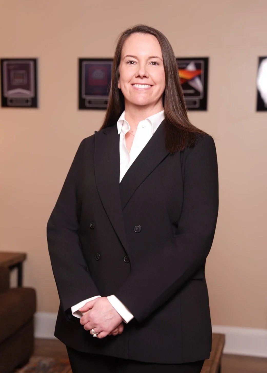 A professional woman with long brown hair smiling, wearing a black blazer over a white shirt, standing in an office or conference room with framed artwork on beige walls.
