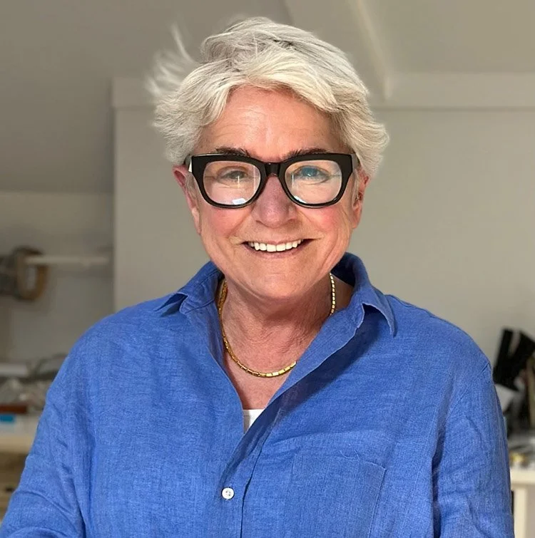 A woman with short, grey, tousled hair wearing black glasses, a blue button-up shirt, and a gold necklace, smiling indoors.
