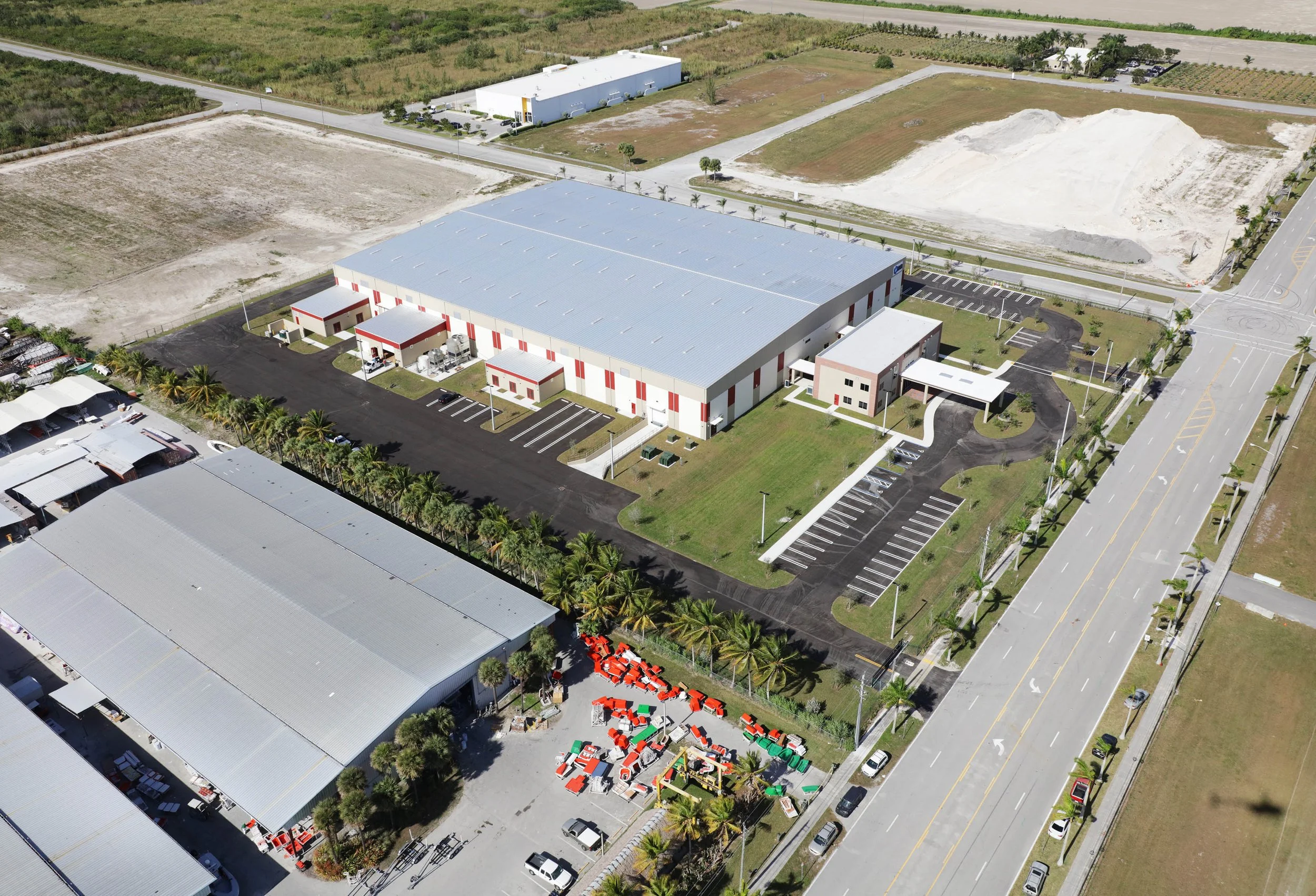 Aerial view of a large industrial warehouse with surrounding paved parking lots, landscaping with palm trees, and adjacent empty lots, with roads and other buildings in a semi-rural area.