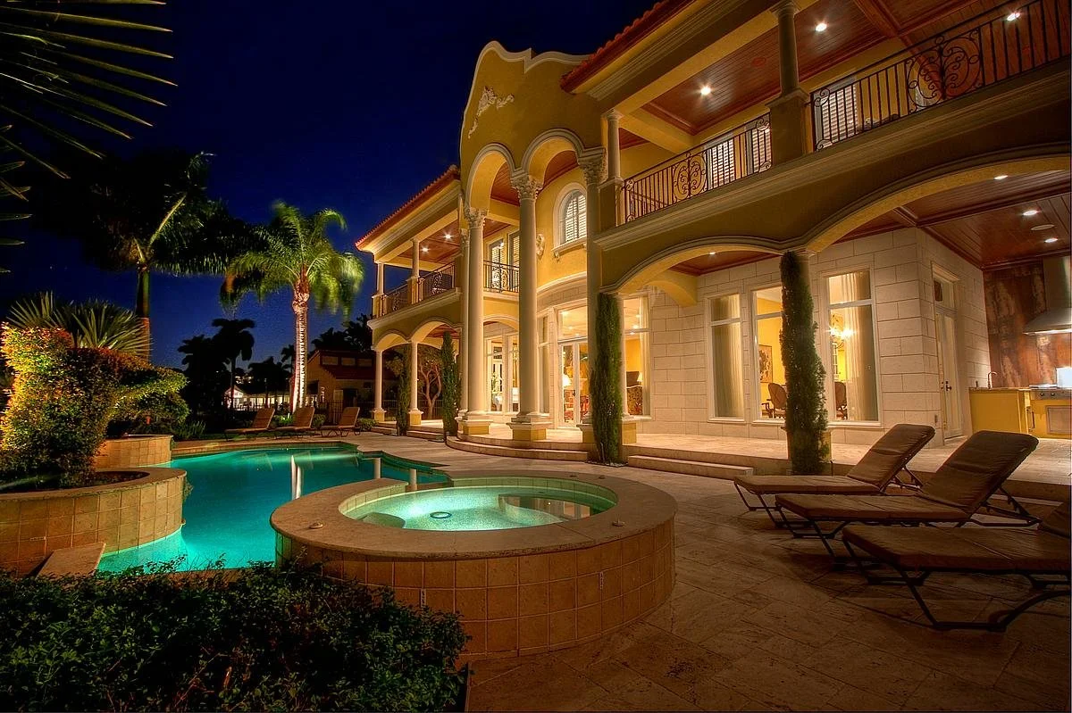 Luxurious backyard with a heated pool and hot tub, surrounded by lounge chairs, palm trees, and illuminated by outdoor lighting at night. The house has large windows, a balcony, and decorative columns.