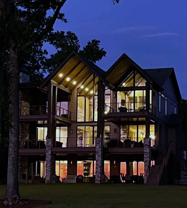 A large, multi-story house illuminated from inside at dusk, with tall windows, balconies, and exterior stairs, surrounded by trees and a lawn.