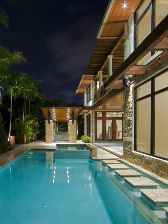 Night view of a modern house with large glass windows and a swimming pool surrounded by lush greenery.