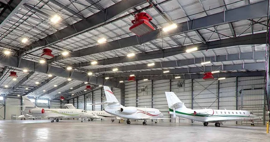 Three private jets parked inside a spacious aircraft hangar with a high ceiling and large lighting fixtures.