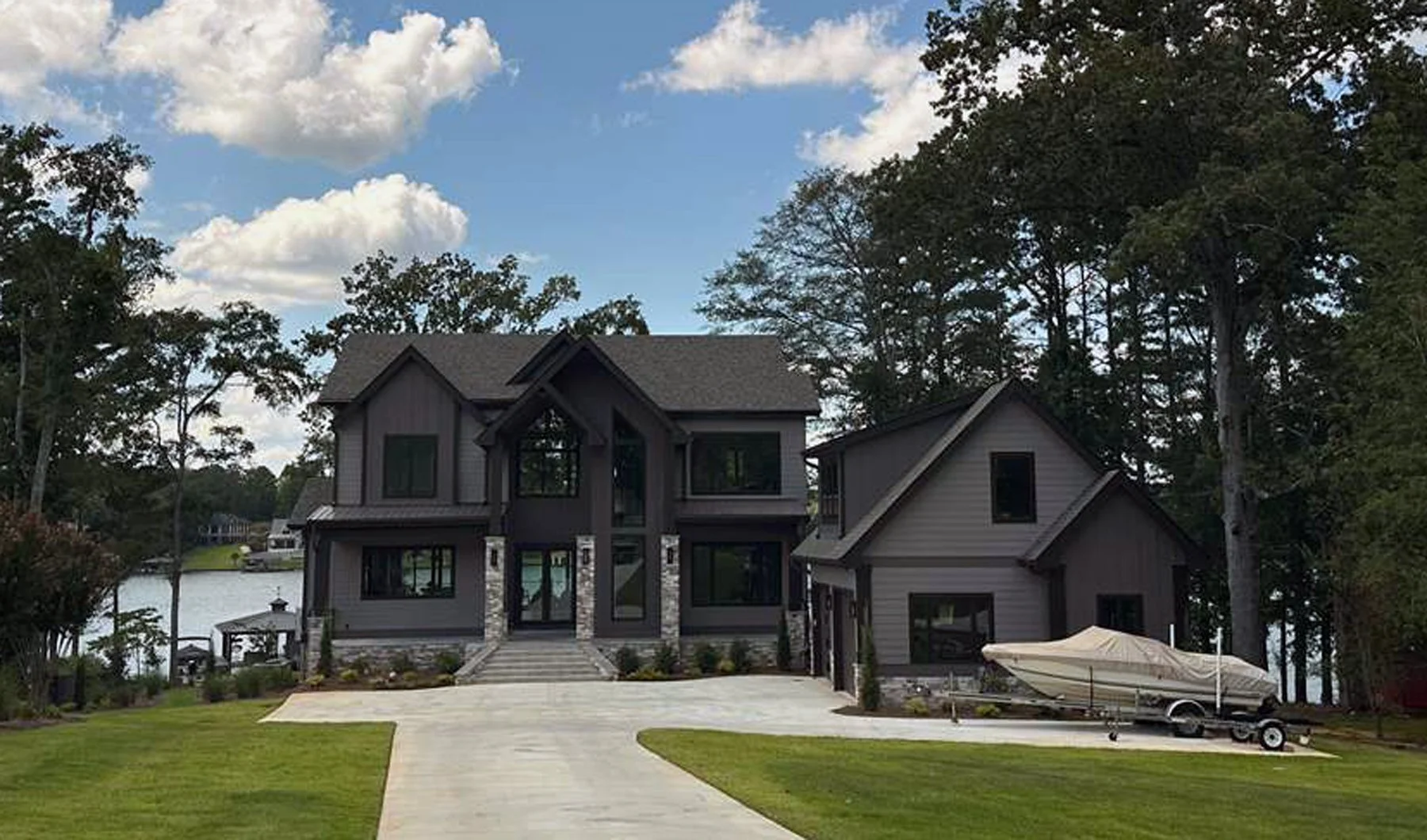 A modern two-story house with gray siding and large windows is situated beside a lake, with trees surrounding it. A boat on a trailer is parked on the driveway near the house, and the lawn is green and well-maintained.