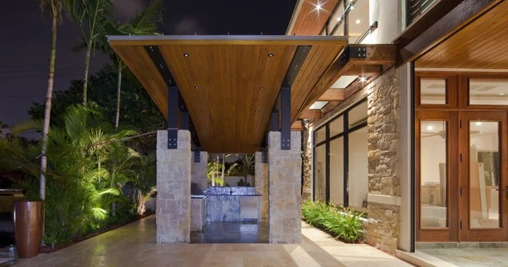 Night view of an outdoor patio with a wooden overhang supported by stone columns, adjacent to a house with large glass doors and modern design elements, surrounded by lush tropical plants.