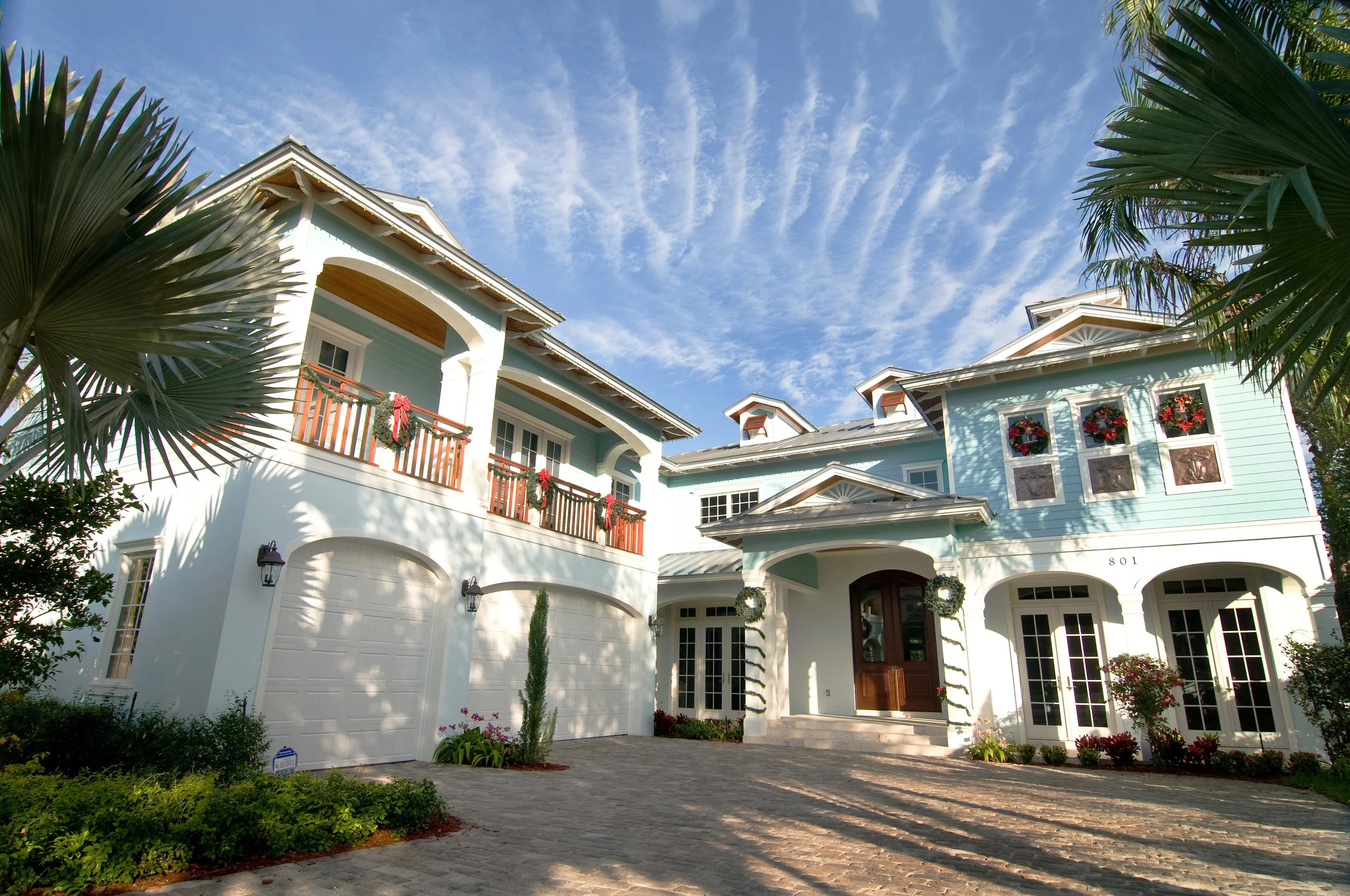 A large, light blue house decorated with holiday wreaths and garlands, featuring a cobblestone driveway and lush tropical plants, under a partly cloudy sky.