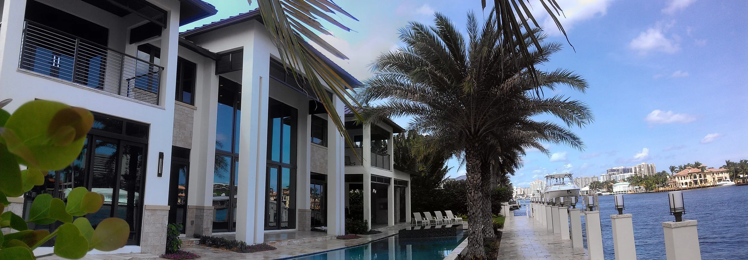 Modern waterfront house with large windows, a balcony, palm trees, and a boat docked nearby along a canal under a partly cloudy sky.