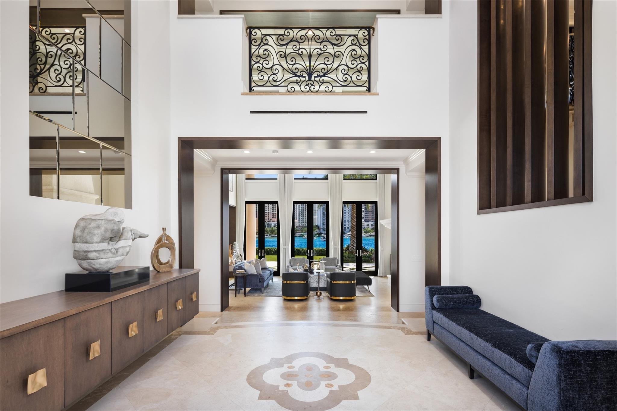 A spacious, modern living room with high ceilings, large windows, and elegant decor. There is a dark wooden sideboard with sculptures on top on the left, a blue upholstered bench on the right, and a view of a lake with city buildings in the backgroun