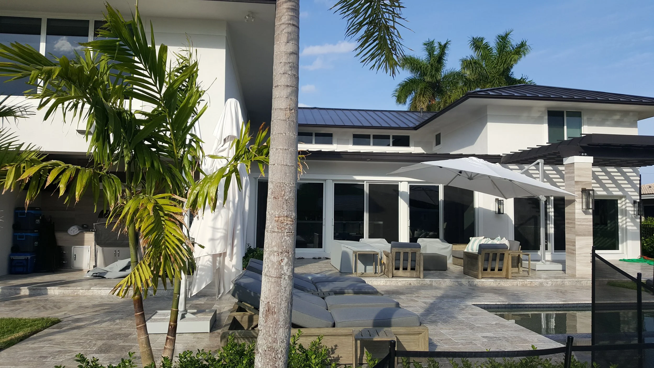 Backyard patio with outdoor furniture, umbrella, palm trees, and a modern house with large glass sliding doors and a metal roof.