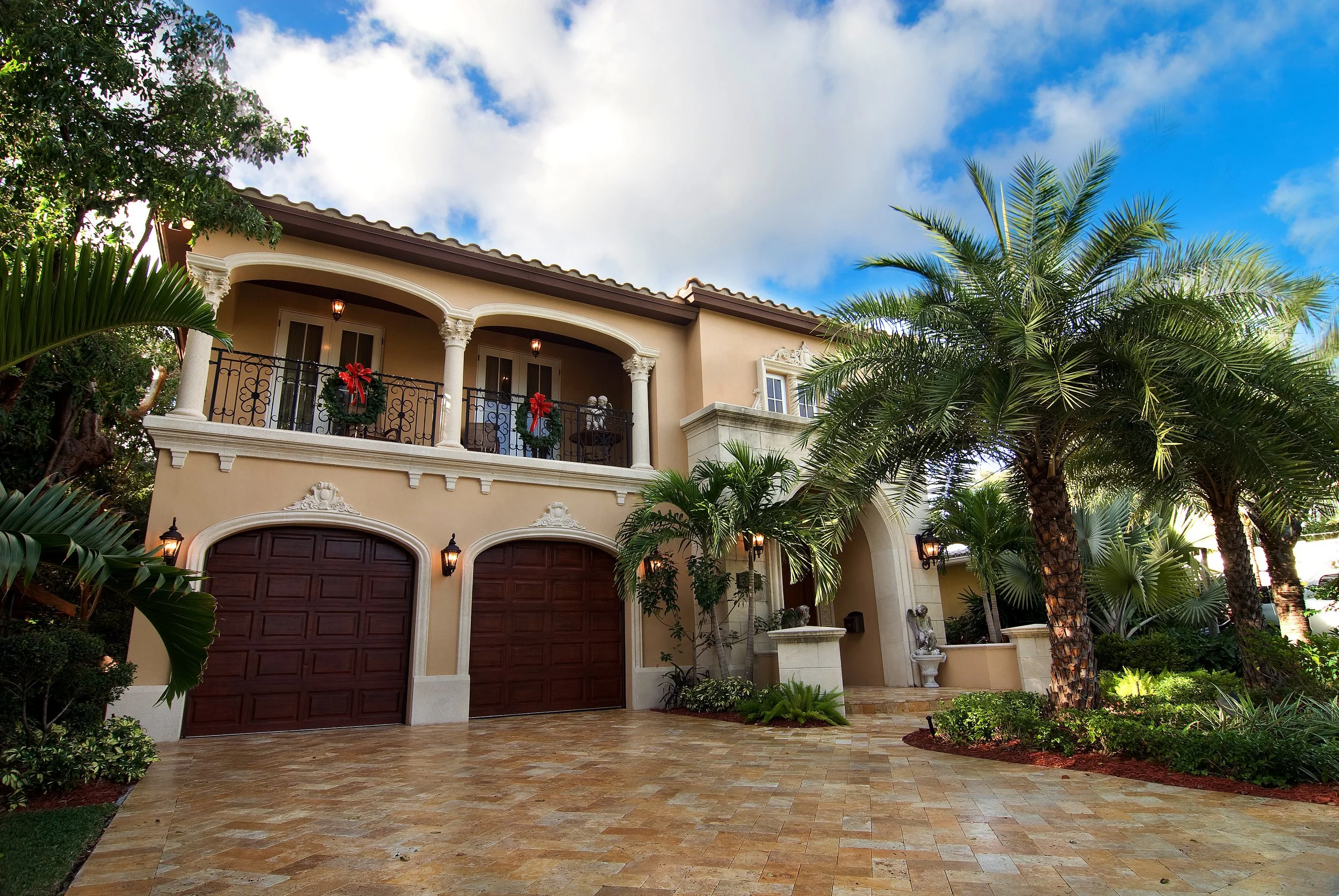A luxurious two-story house with a beige exterior, dark brown garage doors, and decorative wrought-iron balcony railings decorated with Christmas wreaths, surrounded by lush palm trees and tropical landscaping under a partly cloudy sky.
