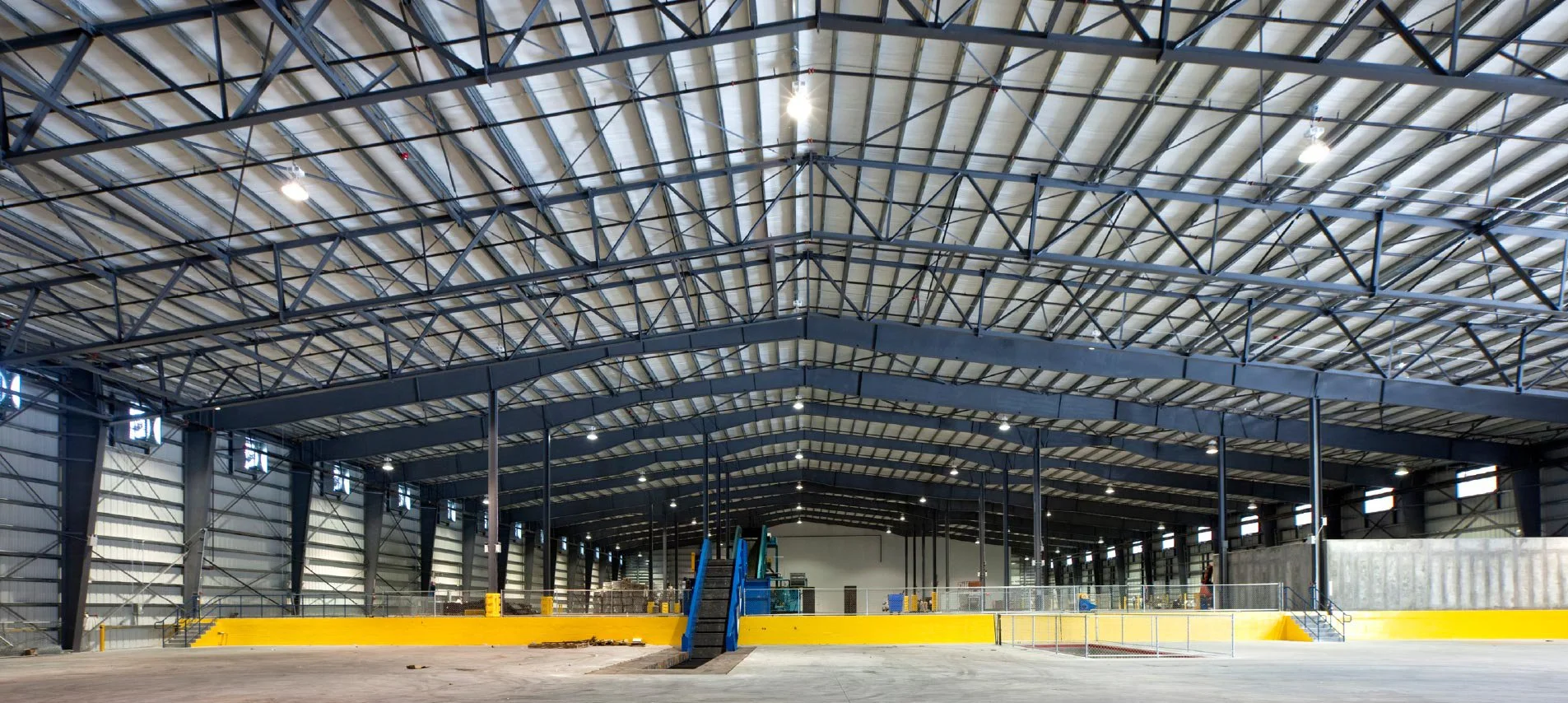 Inside a large industrial warehouse with a high arched metal roof and steel beams, unfinished concrete floor, and some construction equipment and materials, including a blue staircase, yellow safety barriers, and scattered debris.